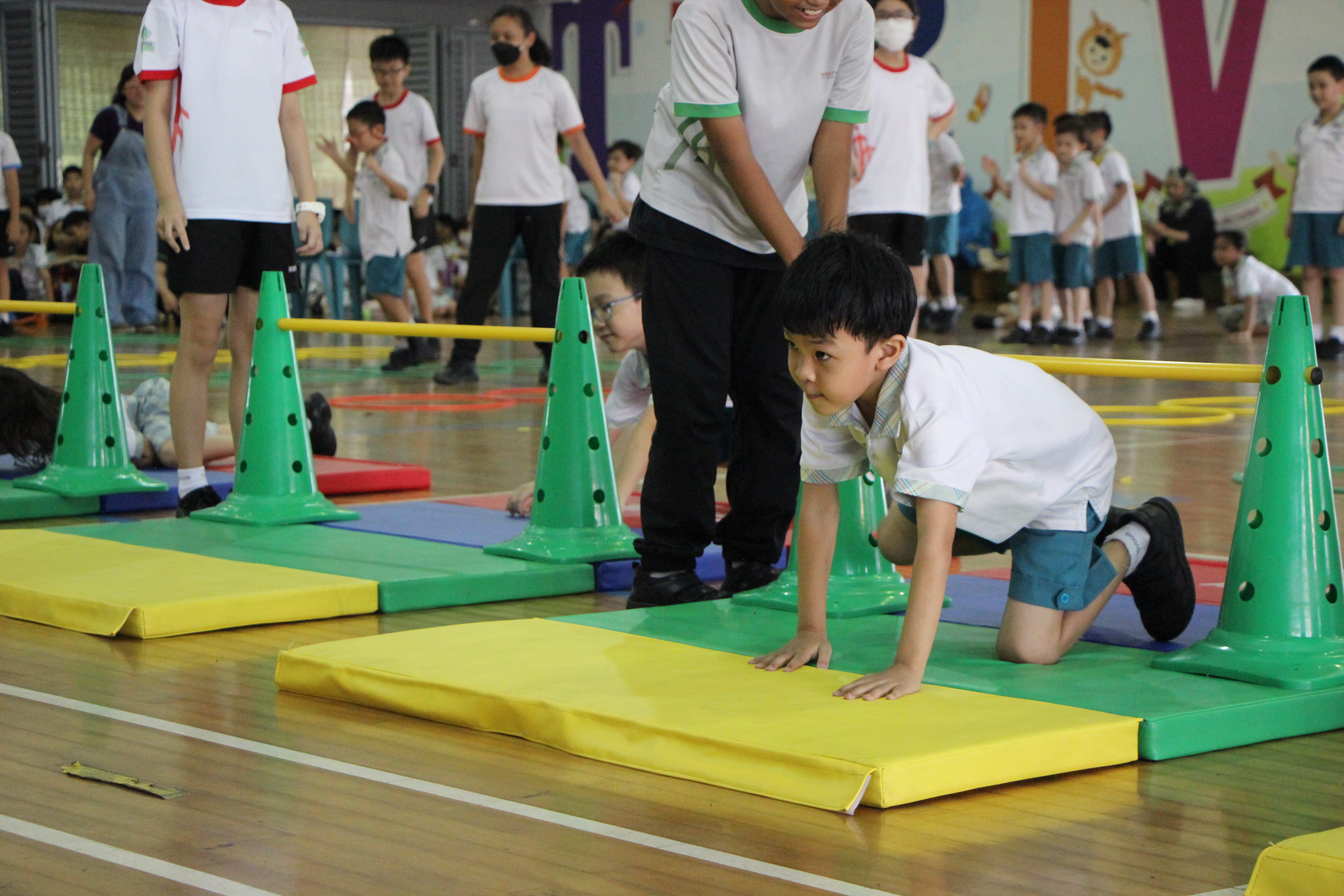 A young boy crawls on colorful foam mats between green cones during an indoor school gym activity.